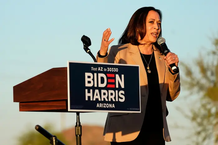 Senator Kamala Harris in a tan jacket, black shirt and pants, holding a microphone by a lectern with a sign that says Biden Harris Arizona. She is outside, with the desert in the background.